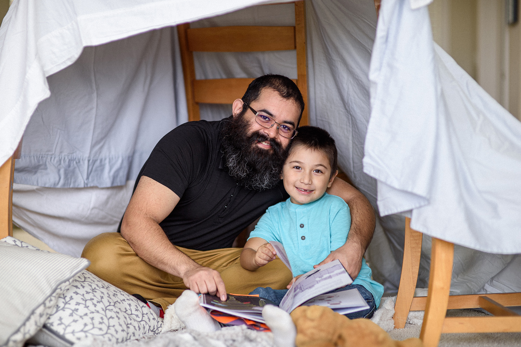 Padre e hijo leyendo libros debajo de un fuerte hecho con una sábana y sillas